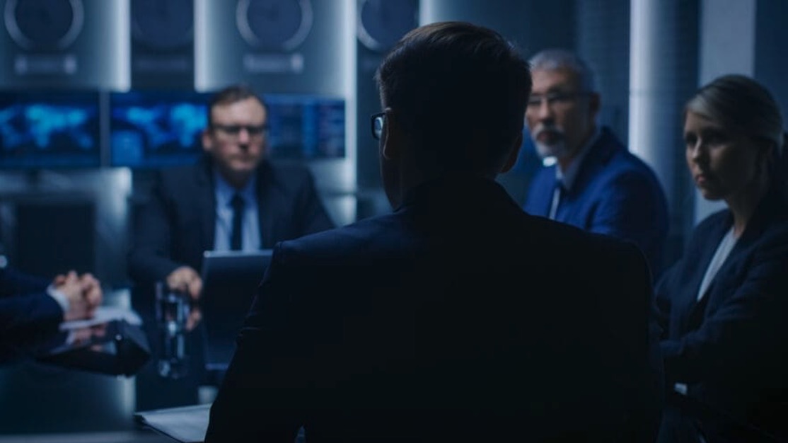 Group of businesspeople in a dimly lit meeting room with multiple screens in the background.