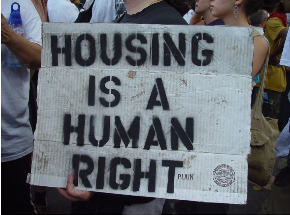 Person holding a cardboard sign reading ‘HOUSING IS A HUMAN RIGHT’ at a protest.