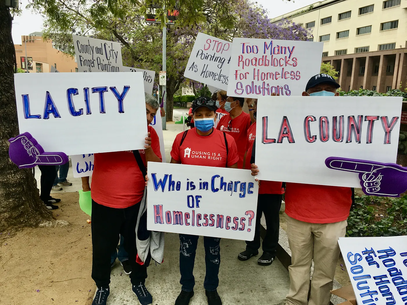 Activists holding signs about homelessness, including ‘LA CITY,’ ‘LA COUNTY,’ and ‘Who is in Charge of Homelessness?